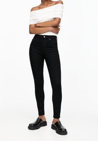 Jeans ajustados de talle alto en color negro, fabricados en denim con una textura suave, combinados con una blusa blanca de hombros descubiertos y zapatos negros gruesos.