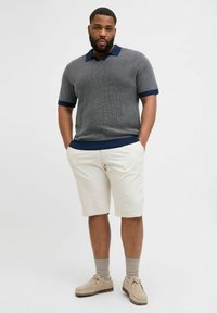 Grå kortærmet polo med en marineblå krave, parret med lyse beige shorts. Modellen har beige sneakers og grå sokker på.
