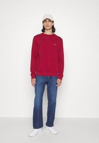 Felpa rossa con finitura testurizzata e un piccolo logo sul petto, abbinata a jeans blu in denim e sneakers bianche, indossata da un modello maschile.