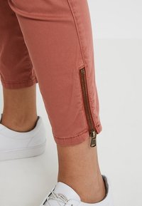 Pantalons cropped couleur rouille en tissu doux, avec un détail de fermeture éclair sur le côté et des ourlets terminés. Associés à des baskets blanches.