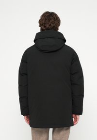 Casaco acolchoado preto com capuz, apresentando uma textura suave e mangas compridas. O design inclui um corte recto e detalhes minimalistas em ferragens.