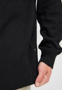 Suéter de punto negro con puños y dobladillo acanalados, que presenta una textura similar a la de un suave rizo, y una pequeña etiqueta en el costado.