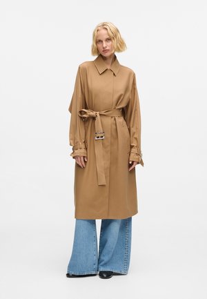 Abrigo trench oversized color beige con cuello, cinturón de hebilla cuadrada y mangas dobladas, llevado sobre unos vaqueros de pierna ancha azules y botas negras.