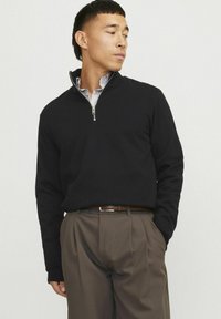 Melnā zip-up jaka ar garām piedurknēm, ribotām aprocēm un sudraba rāvējslēdzēju. Nēsāta virs svītraina krekla ar apkakli un apvienota ar brūniem biksēm.