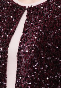 Bordo aksomo audinys, tankiai padengtas tamsiai violetiniais blizgučiais, matomas po balta medžiaga apykaklėje.