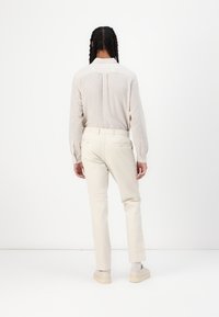 Camisa de manga larga beige claro con un canesú en la parte posterior, combinada con pantalones ajustados color crema y zapatos beige claro, sobre un fondo blanco.