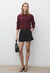 Burgunderröd kort jacka med guldknappar, framfickor, kombinerad med svarta veckade shorts och svarta flip-flops, håller en svart handväska.