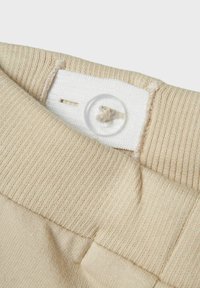 Beige tyg med ribbad textur, med en vit etikett fäst vid en genomskinlig knapp. Synlig sömdetalj nära etiketten.