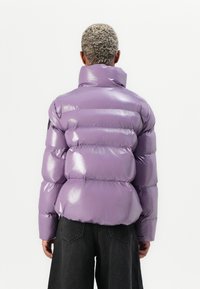 Lilla pufferjakke med en blank overflade, høj krave og vandret syninger. Har et sort logo-patch på ærmet.