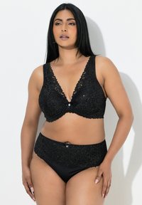 Kvinde med langt sort hår iført en matchende sort blonder-bh og højtaljede underbukser, stående mod en ensartet hvid baggrund.