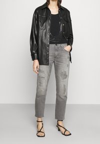Camisa chaqueta de piel sintética negra con botones dorados, llevada sobre una camiseta negra sin mangas con un diseño brillante. Jeans desgastados de color gris claro y sandalias negras con tiras.