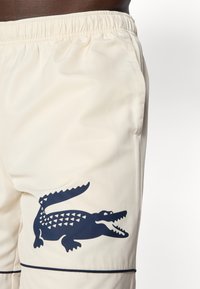 Cream-colored shorts featuring a textured fabric, elastic waistband, and a dark blue crocodile logo with geometric details.