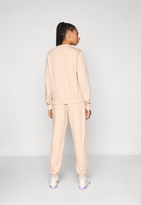 Gaiši bēšs džemperis un atbilstošas bikses no mīksta auduma. Apģērbam ir ribotas aproces un brīvs piegriezums, valkātas ar baltām sporta kurpēm.