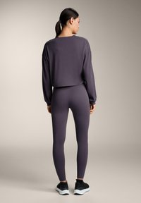 Ljubičasti kratki dukserica s dugim rukavima, uparen s uskim tajicama istog uzorka. Model nosi crne atletske tenisice. Glatka tekstura, minimalni dizajn.