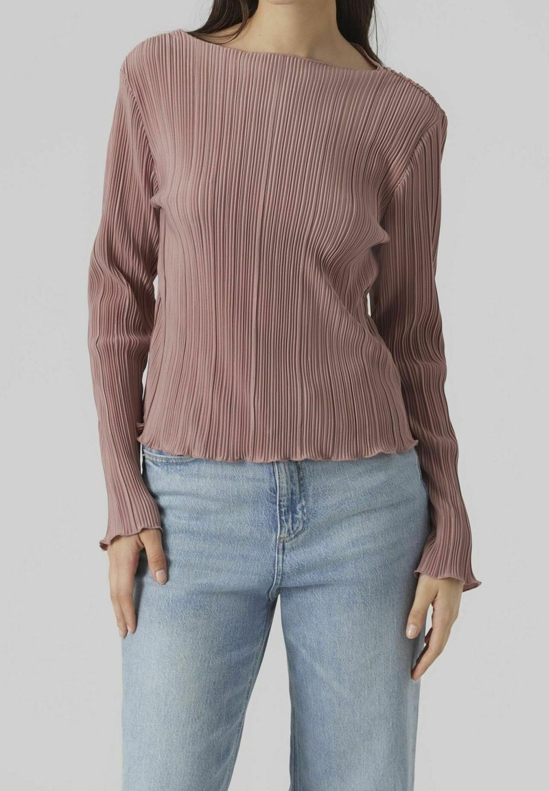 Dlouhý rukáv, řasený top v barvě pudrové růže, s lodičkovým výstřihem a třásněmi na lemu, spárovaný s světlými modrými džíny.