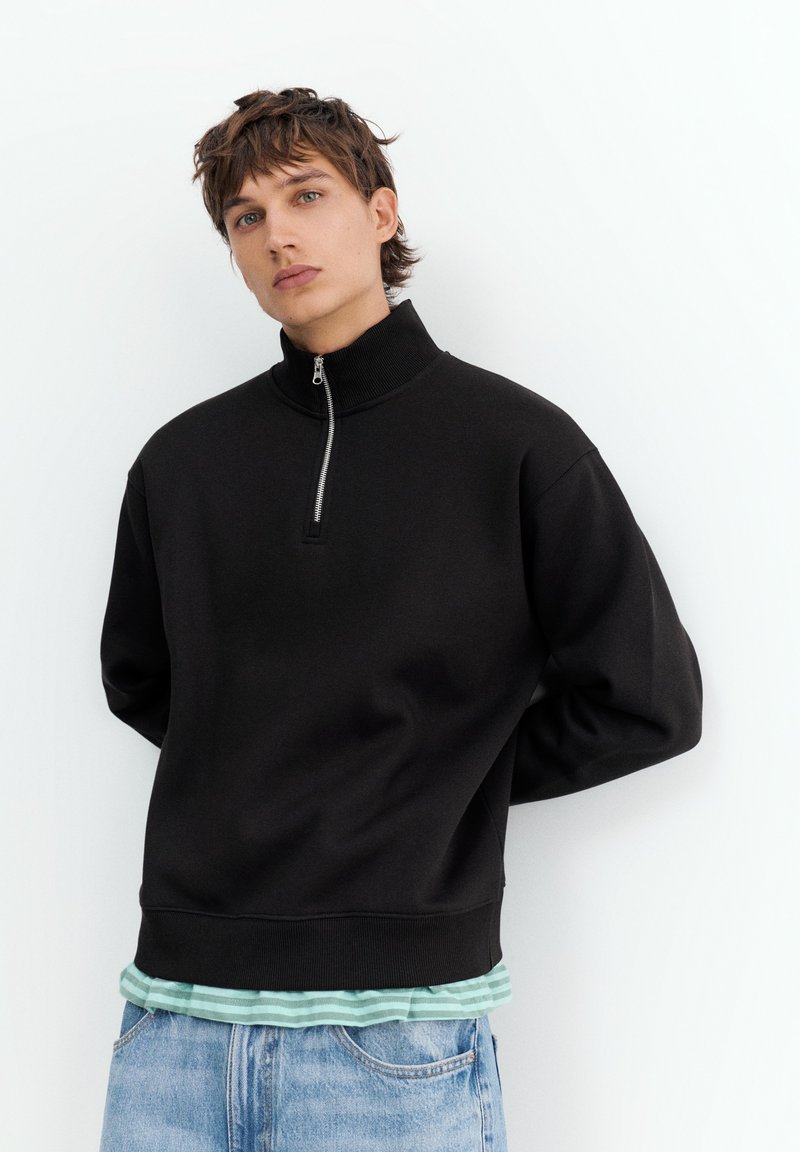 Jeune personne aux cheveux courts et bruns portant un pull noir zippé sur une chemise rayée verte et un jean bleu clair sur fond blanc.