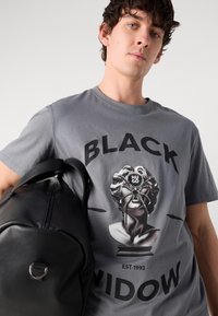 Grå bomullströja med stor svart text "BLACK WIDOW" och en grafisk bild av ett skulpterat huvud prytt med en spindel. Svart läder väska.