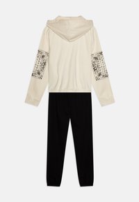 Felpa con cappuccio color crema con stampa floreale nera sulle maniche, abbinata a pantaloni jogger neri. Tessuto morbido, polsini e vita elasticizzati.