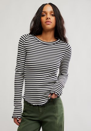 Jeune femme aux longs cheveux tressés portant un t-shirt à manches longues rayé noir et blanc et un pantalon vert, debout devant un fond uni.