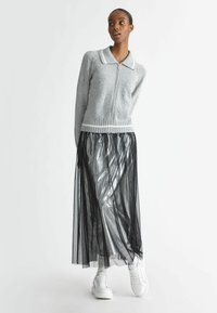 Cardigan grigio con zip, colletto bianco e orlo a costine abbinato a una gonna nera fluida e trasparente e scarpe da ginnastica bianche, che mettono in mostra strati di texture.