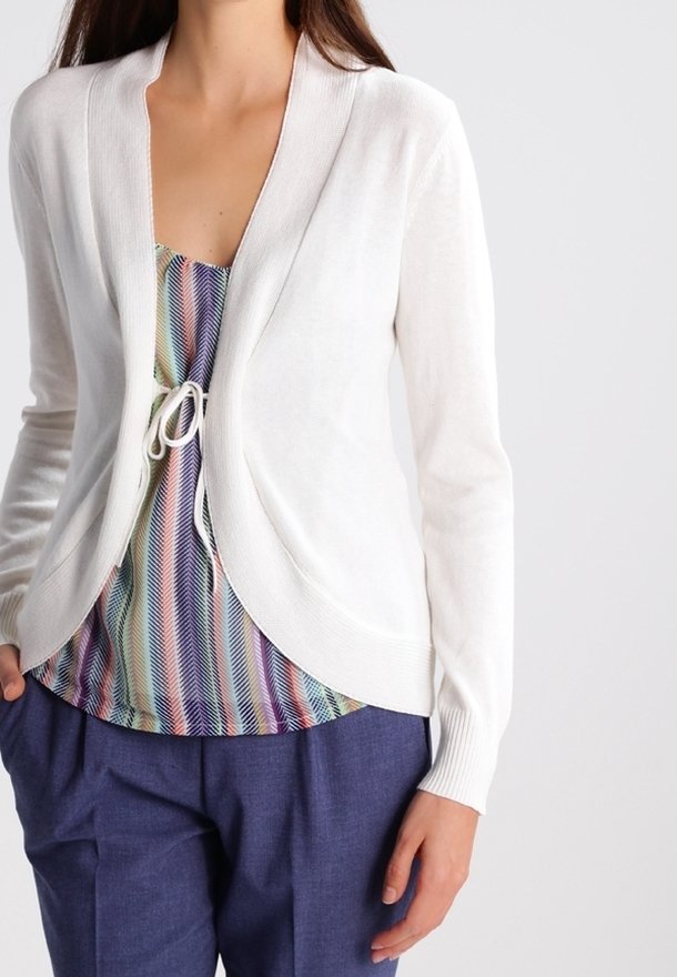 Cárdigan blanco, ligero, con manga abierta y puños acanalados. Lleva puesto un top de rayas multicolores y está combinado con pantalones oscuros.