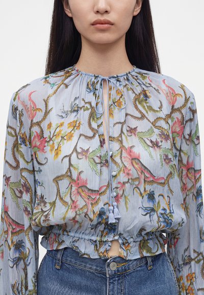Femme portant un chemisier bleu clair à motifs floraux avec un cordon à l’encolure, associé à un jean bleu, vue des épaules à la taille.
