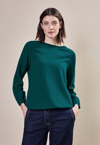 Tealblus med texturerad yta, långa ärmar och volangkrage, kombinerad med mörka jeans och en neutral bakgrund.