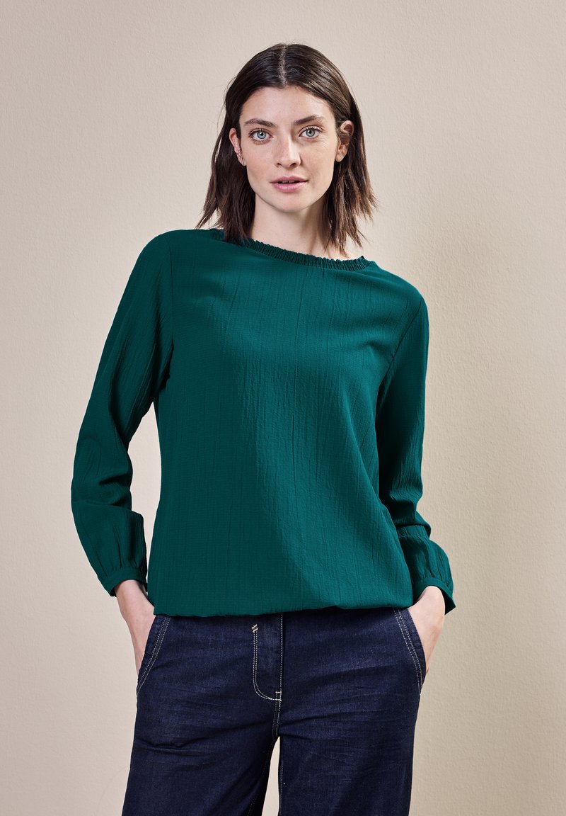 Tealblus med texturerad yta, långa ärmar och volangkrage, kombinerad med mörka jeans och en neutral bakgrund.
