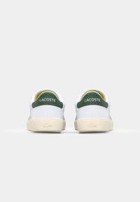 White sneakers with green accents, featuring a smooth leather upper, round toe, padded collar, and beige rubber sole. LACOSTE logo visible.