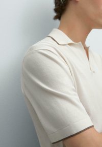 Polo shirt crema con colletto e maniche corte. Presenta un tessuto testurizzato e rifiniture a contrasto ai polsini, che conferiscono un tocco di dettaglio discreto.