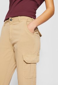 Beige cargo bukser med sidelommer, broderte detaljer og en avslappet passform. Laget av myk stoff, med knappeåpning og beltehemper.