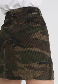 Jupe mini en velours côtelé avec un imprimé camouflage, présentant des nuances de vert et de marron, avec une taille ajustée et un tissu texturé.