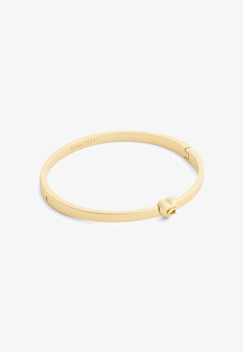 Bracelet jonc en métal doré Coach avec un fermoir en forme de cœur et un logo gravé sur la face intérieure.