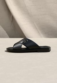 Μαύρο δερμάτινο σανδάλι τύπου slide με λουριά που διασταυρώνονται και μεταλλικό έμβλημα, τοποθετημένο πάνω σε ανοιχτό μπεζ φόντο με υφή.