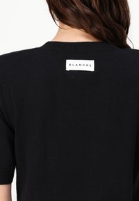 Schwarzer Strickpullover mit kurzen Ärmeln und Rippkragen, von hinten gezeigt. Besitzt ein weißes Etikett am Kragen mit der Aufschrift "BLANCHE".