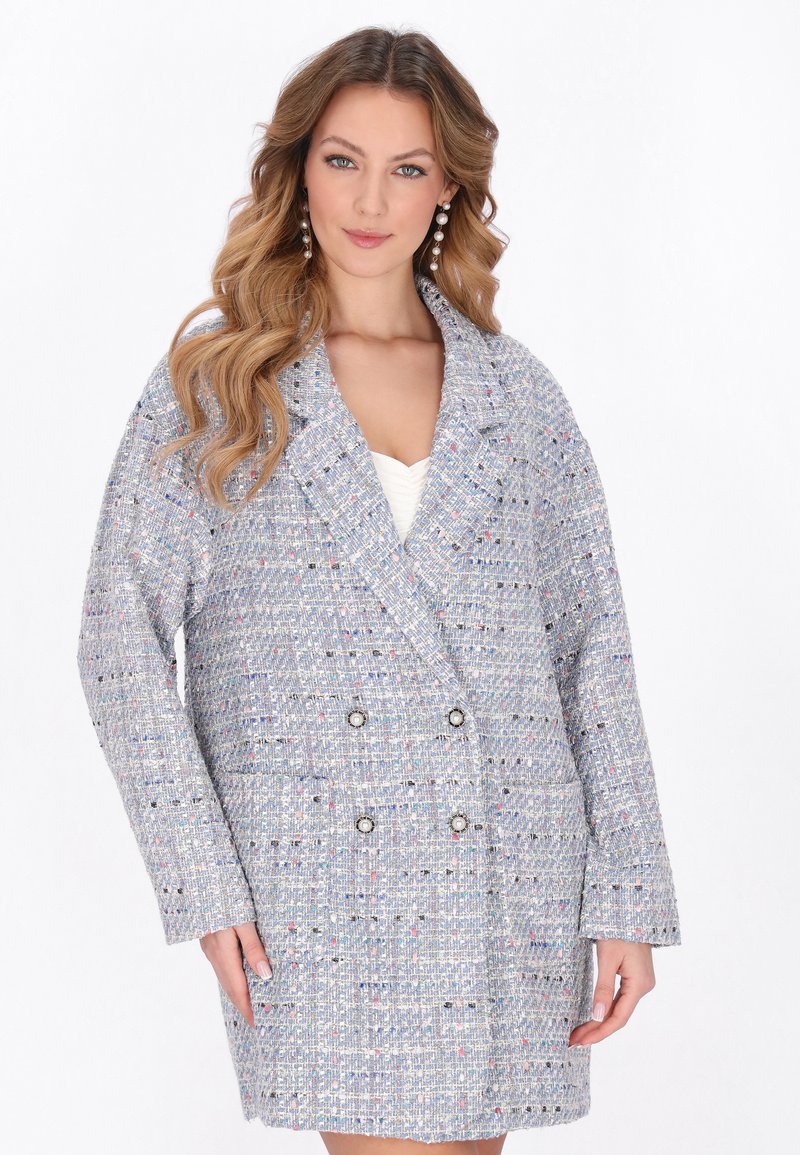 Donna che indossa un cappotto doppiopetto in tweed azzurro chiaro e multicolore con bottoni di perla e orecchini pendenti di perla, in piedi su sfondo bianco.