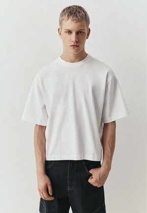 Ung mand med kort blondt hår iført en almindelig, hvid, oversized T-shirt og mørke jeans, der står op ad en hvid væg.