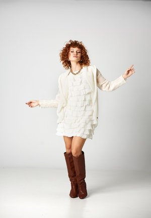 Kobieta z kręconymi rudymi włosami, ubrana w białą falbanianą sukienkę, kremowy sweter, złoty naszyjnik i brązowe kozaki za kolano, stojąca z wyciągniętymi ramionami.