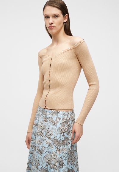 Γυναίκα που φοράει μπεζ πουλόβερ με κουμπιά, ριγωτό και με ανοιχτούς ώμους, και φούστα ανοιχτού μπλε χρώματος με καφέ τροπικό φυτικό σχέδιο.