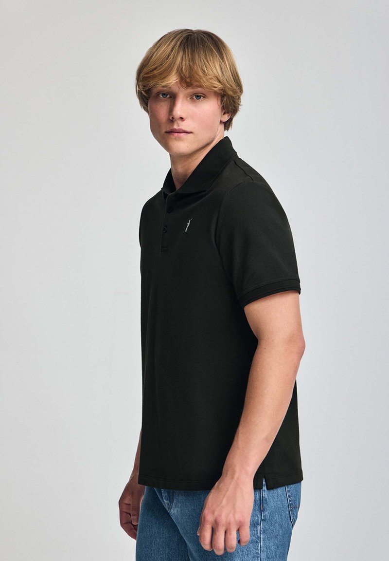 Svart pikétröja med krage, med en liten logotyp på bröstet, ihop med ljusblå jeans, visad från sidan.