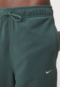 Gröna fleecebyxor med elastisk midjeband och dragsko. Har en liten vit Nike-logotyp på vänster höft. Mjuk textur.