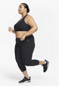 Svart sports-bh og leggings laget av et glatt stoff, med en tettsittende design og Nike-logoer. Modellen jogger i svarte joggesko.