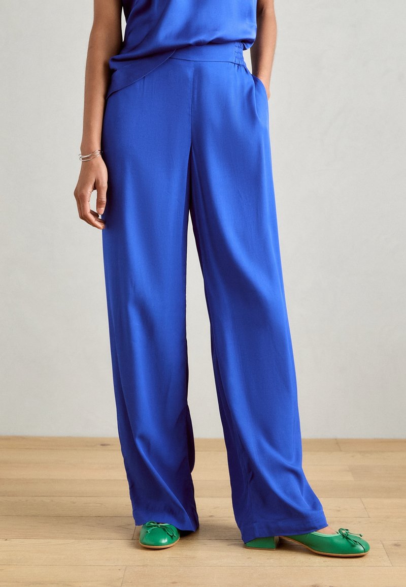 Blauwe wijde broek van gladde stof, voorzien van een elastische tailleband en zijzakken, gecombineerd met groene platte schoenen met strikaccenten.