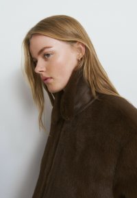 Abrigo de piel marrón con cuello alto, textura suave y diseño sencillo. El perfil de la modelo muestra rasgos suaves y cabello largo.