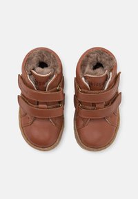 Bottines en cuir marron avec deux straps Velcro et doublure douce et texturée. Bout arrondi et semelle en caoutchouc avec un léger relief pour l'adhérence.