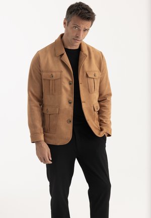 Homme portant une veste beige à boutons avec des poches à rabat et un pantalon noir, debout avec une main dans la poche sur fond blanc.