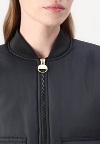 Μαύρο μπουφάν bomber με ομαλή υφή, κλείσιμο με φερμουάρ και στρογγυλό γιακά. Διαθέτει χρυσό τράβηγμα φερμουάρ και πλαϊνές τσέπες.