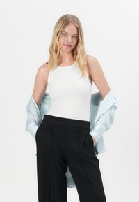 Witte geribde tanktop gecombineerd met hoge zwarte broek en een lichtblauwe overhemd over de schouders gedrapeerd.