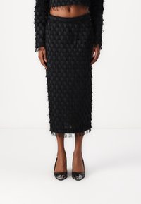 Black textured two-piece outfit featuring a cropped top with fringe details and a midi skirt with a similar pattern. Paired with shiny black heels.