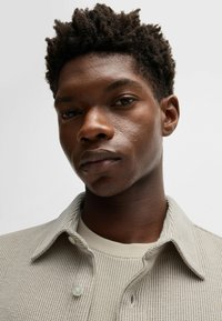 Chemise texturée gris clair avec un col et des détails de boutons, portée par-dessus un haut beige à col rond. Teint de peau lisse et coiffure courte en torsades.